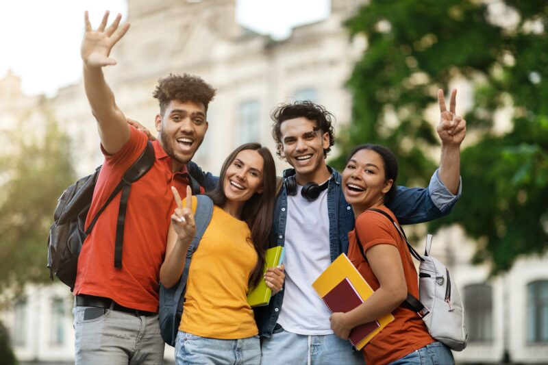 Gruppe fröhlicher Studierender mit Rucksäcken und Büchern posiert lachend vor einem Universitätsgebäude – symbolisiert Freundschaft, Studienanfang und internationales Campusleben.
