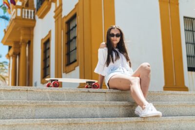 Junge Studentin in sommerlicher Kleidung sitzt mit Sonnenbrille auf einer Treppe neben einem Longboard.