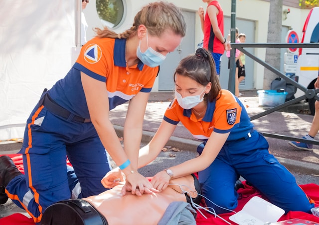 Zwei Rettungssanitäterinnen in Uniform und mit Masken üben Wiederbelebungsmaßnahmen an einer Trainingspuppe im Freien.